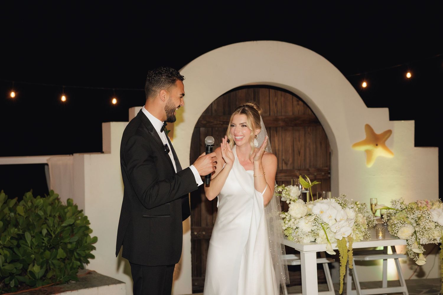 A wedding celebration at dusk with string lights, a white arch, and floral arrangements creating an elegant outdoor setting.