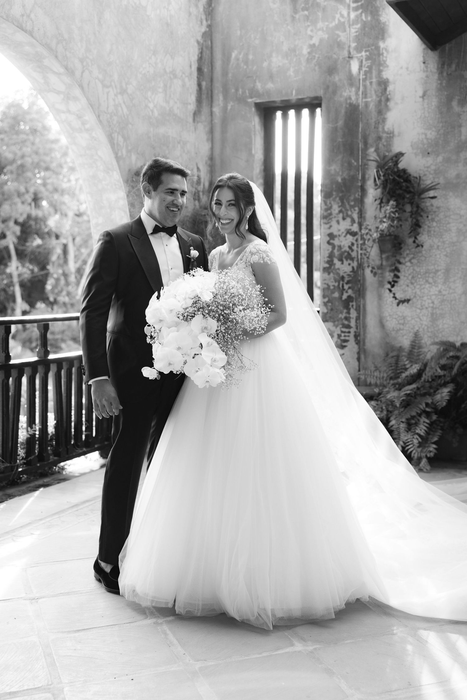 Classic black and white wedding portrait features elegant bridal gown and formal attire against stone archway.