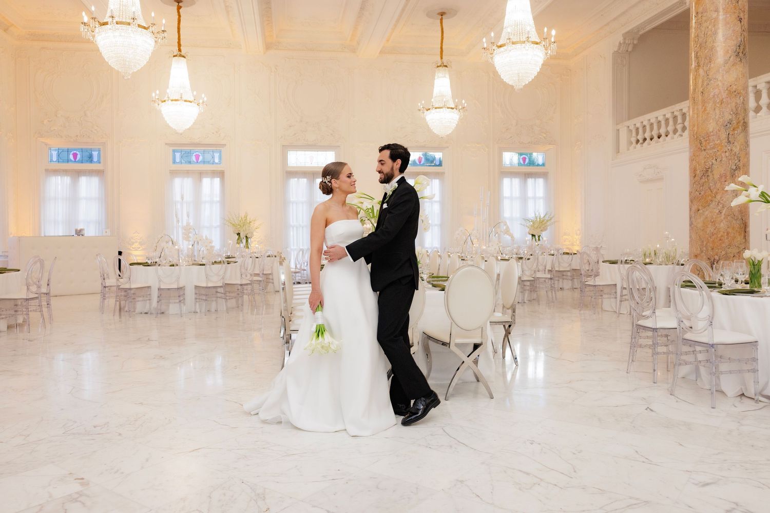 A newly married couple dances together in an elegant white ballroom with crystal chandeliers overhead.