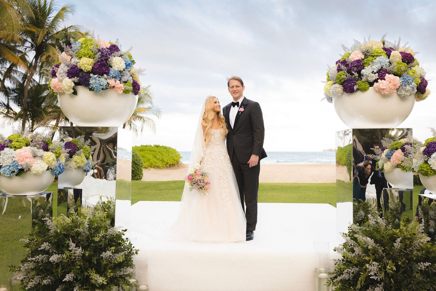 Outdoor wedding ceremony setup with tropical palm trees and elegant floral arrangements around a sunset beach location.