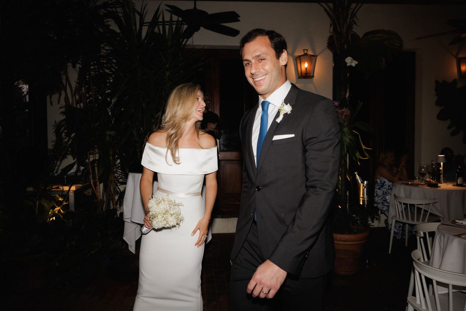 Elegant couple in formal attire smiles together at an evening wedding reception with warm lighting and table settings visible.