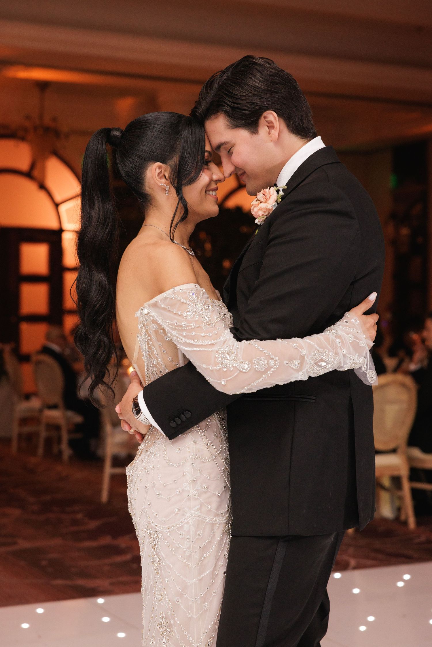 A couple shares their first dance as newlyweds at a wedding reception with romantic lighting and a white dance floor.