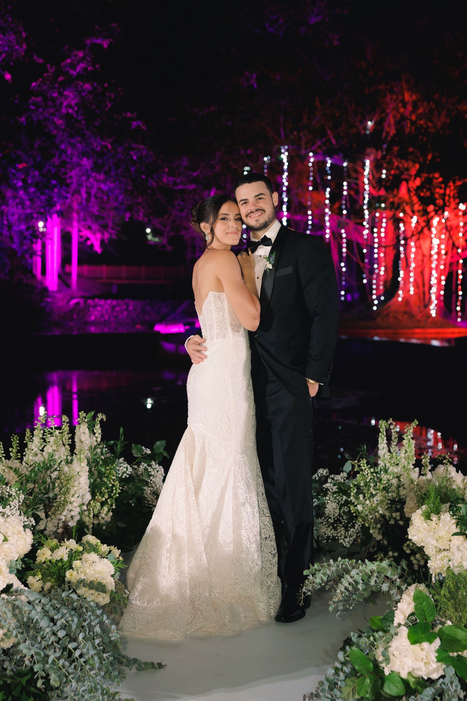 A romantic wedding photo with a couple in formal attire standing in a garden with purple and red lighting.