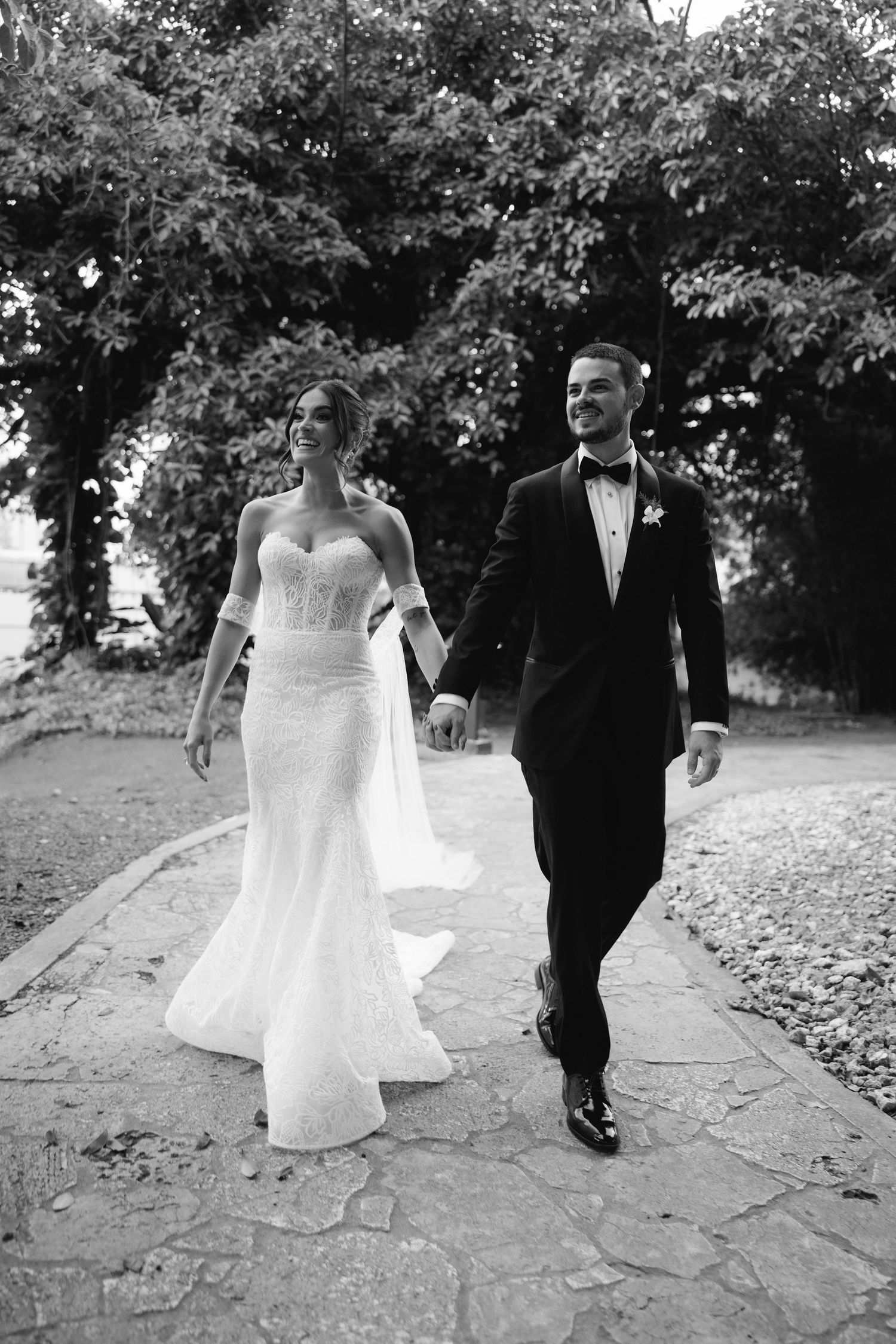 A couple in wedding attire walks through fallen autumn leaves on a stone path in an outdoor setting.