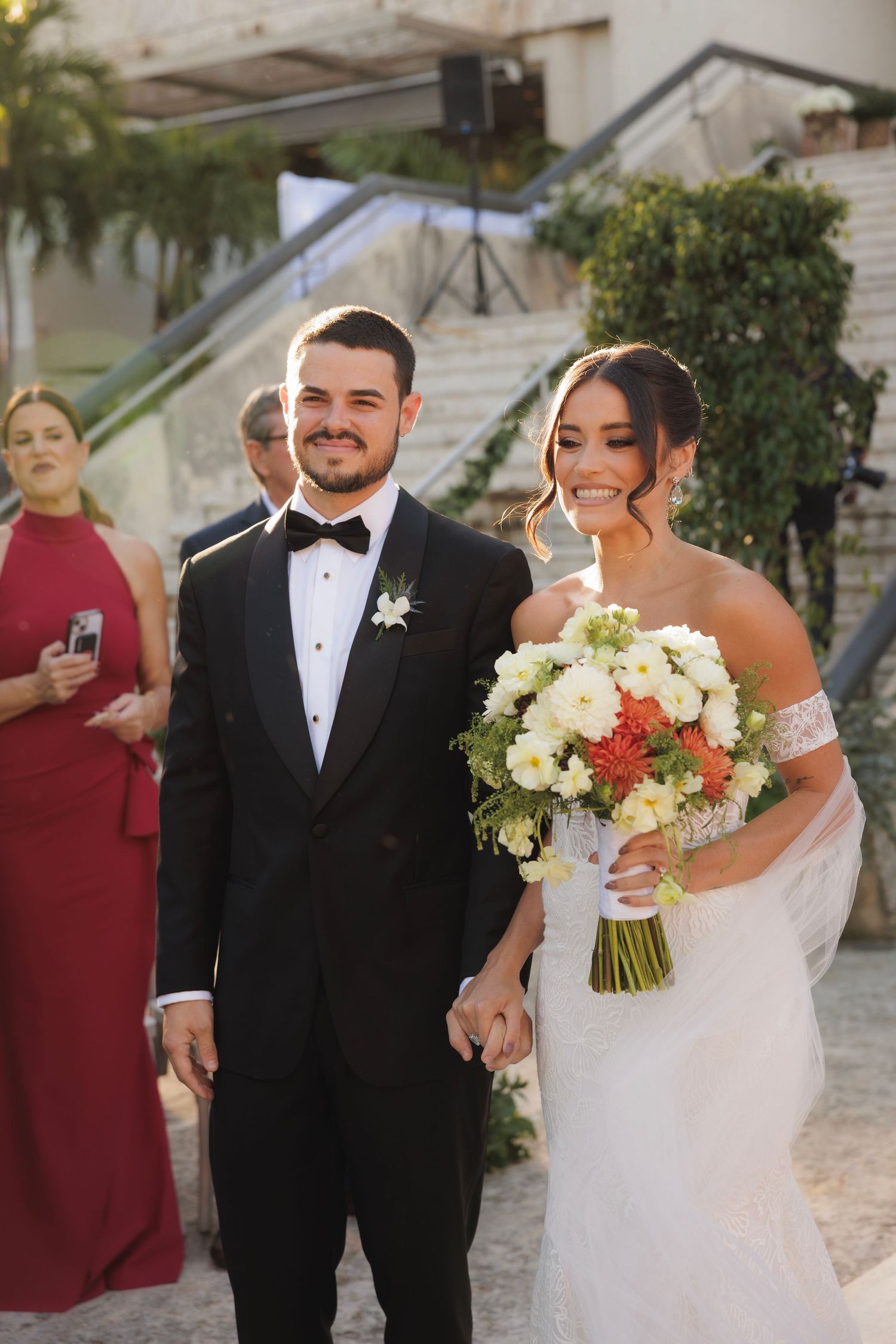 A newlywed couple poses with a white floral bouquet against a stone architectural backdrop.