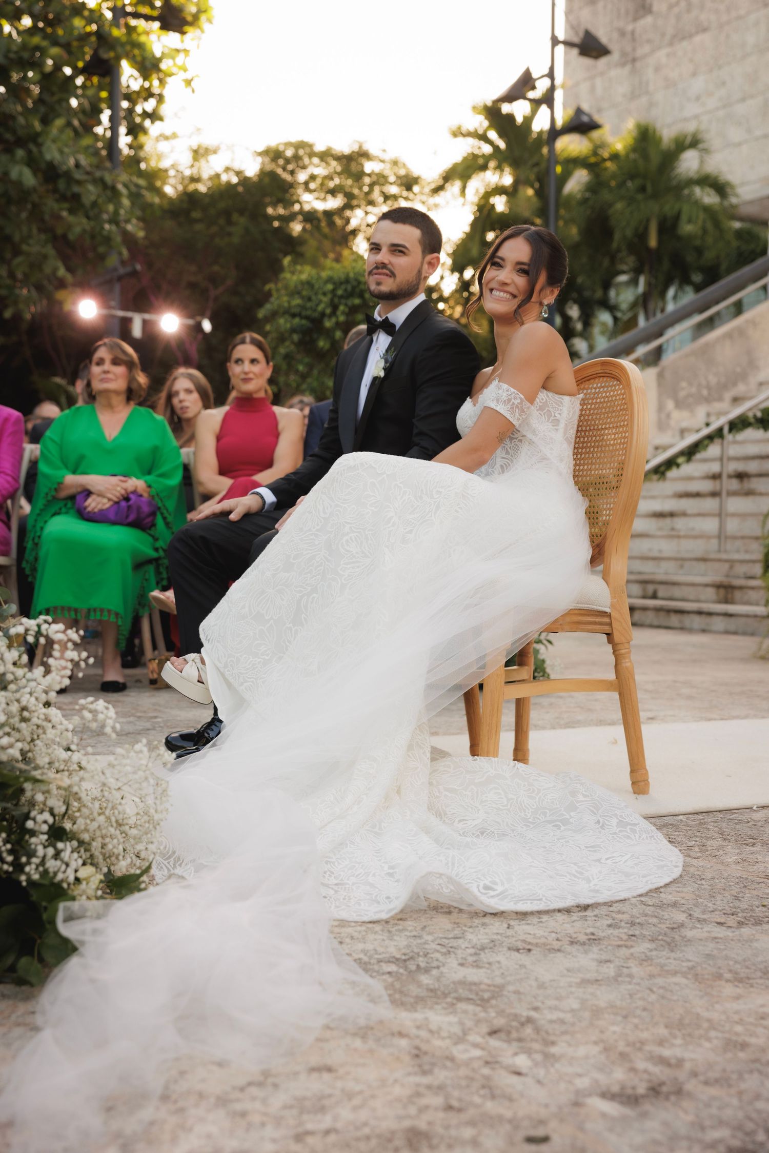 Wedding couple seated on wooden chairs with flowing white dress on stone patio.