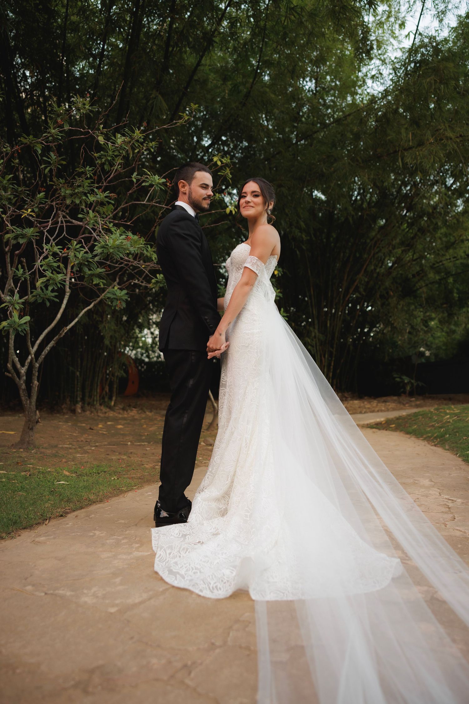 A romantic wedding couple walks along a tree-lined dirt path in an outdoor setting during their photo session.