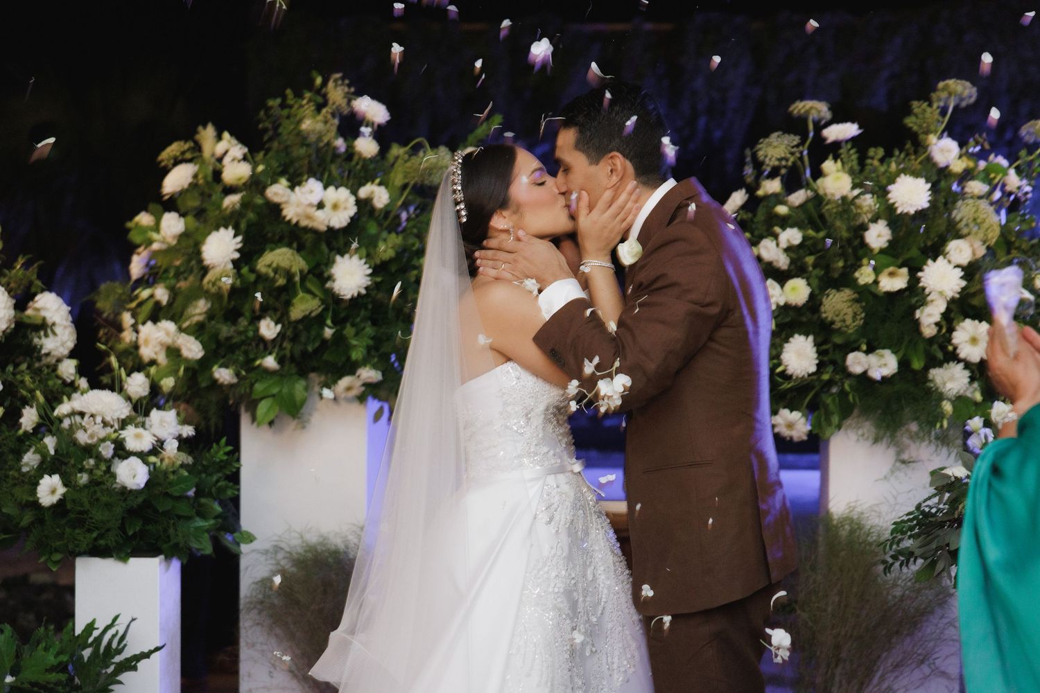 Newlyweds share their first kiss under an elegant floral arch with white roses and greenery.