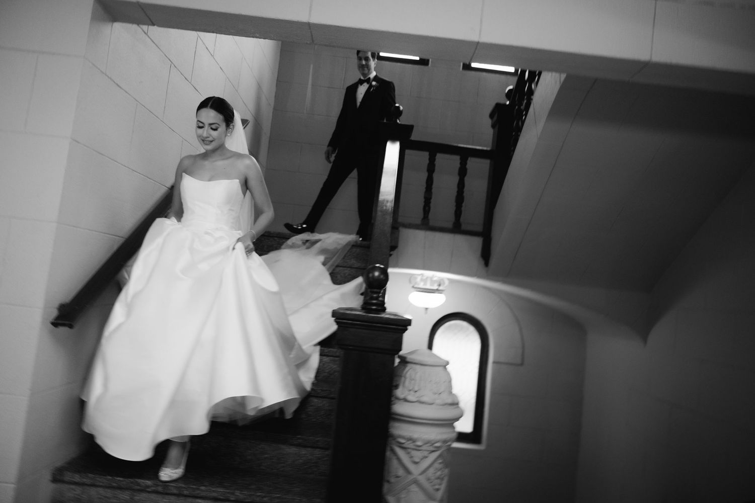 Black and white photograph of a bride in a full wedding gown descending a staircase with others in formal attire.