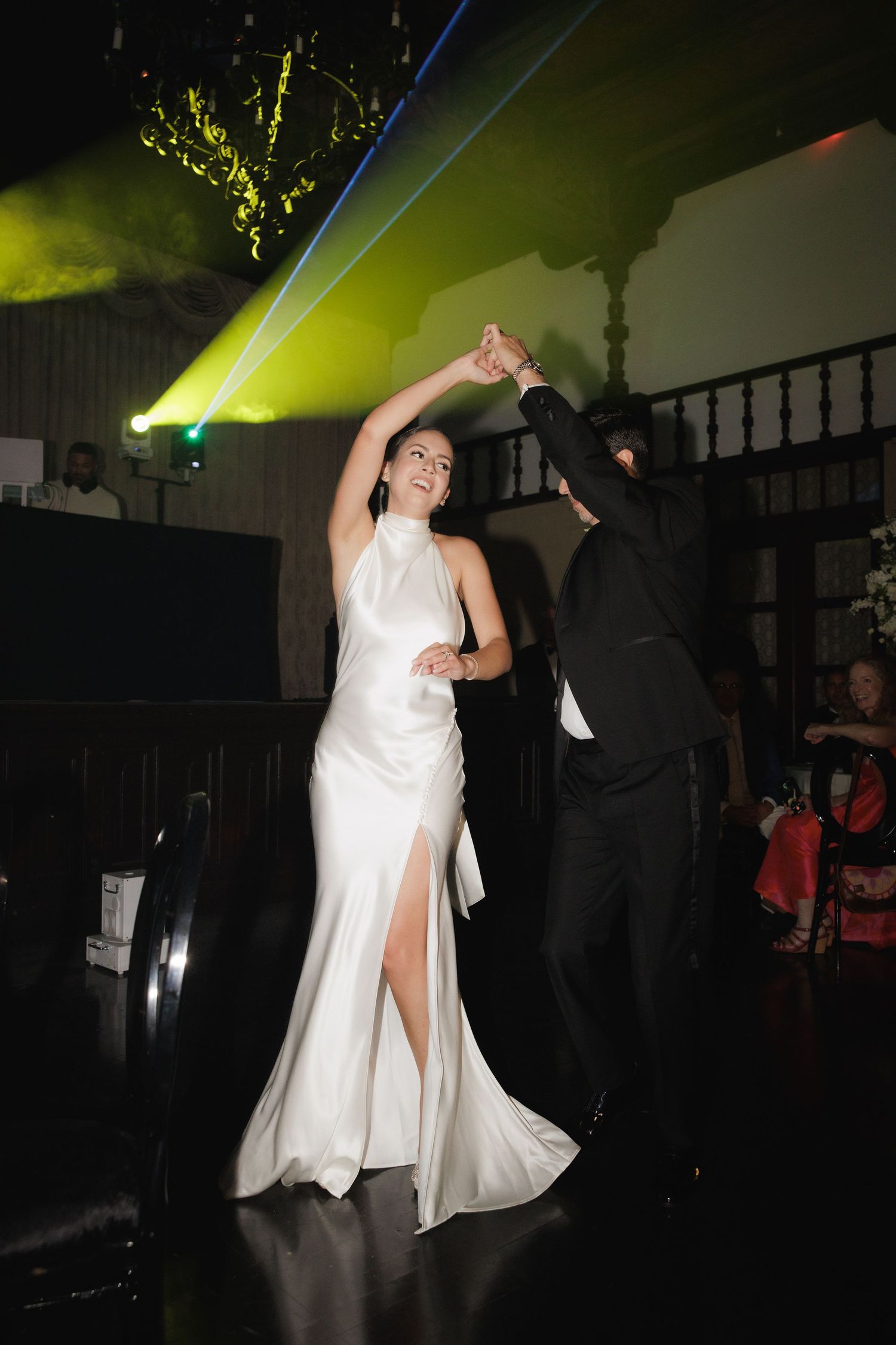 A person in a white dress dances energetically under colorful lighting at what appears to be a wedding reception or party.