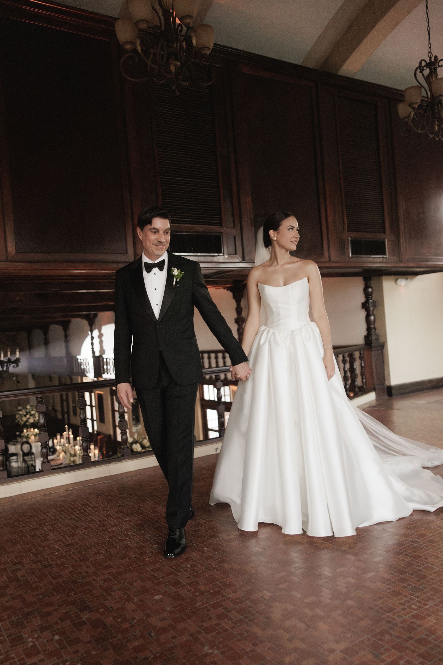 A couple in formal wedding attire walks through an elegant hallway with wooden details and chandeliers.