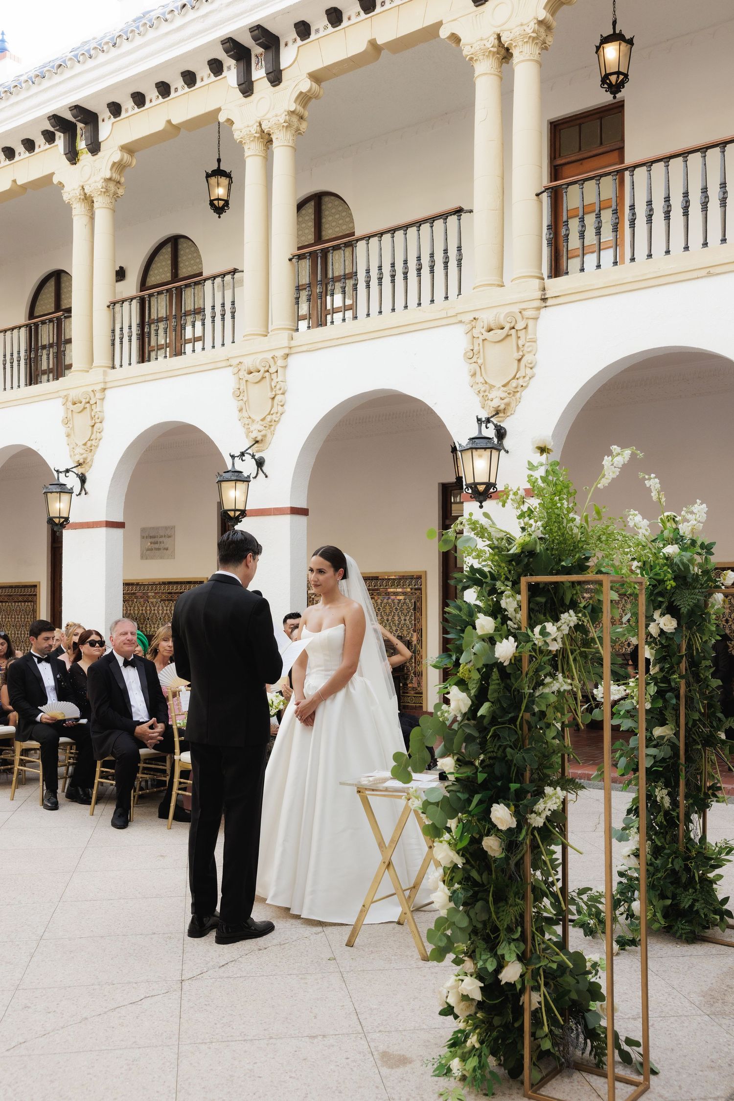 Wedding ceremony setup in an elegant Spanish-style courtyard with white arches and floral decorations.