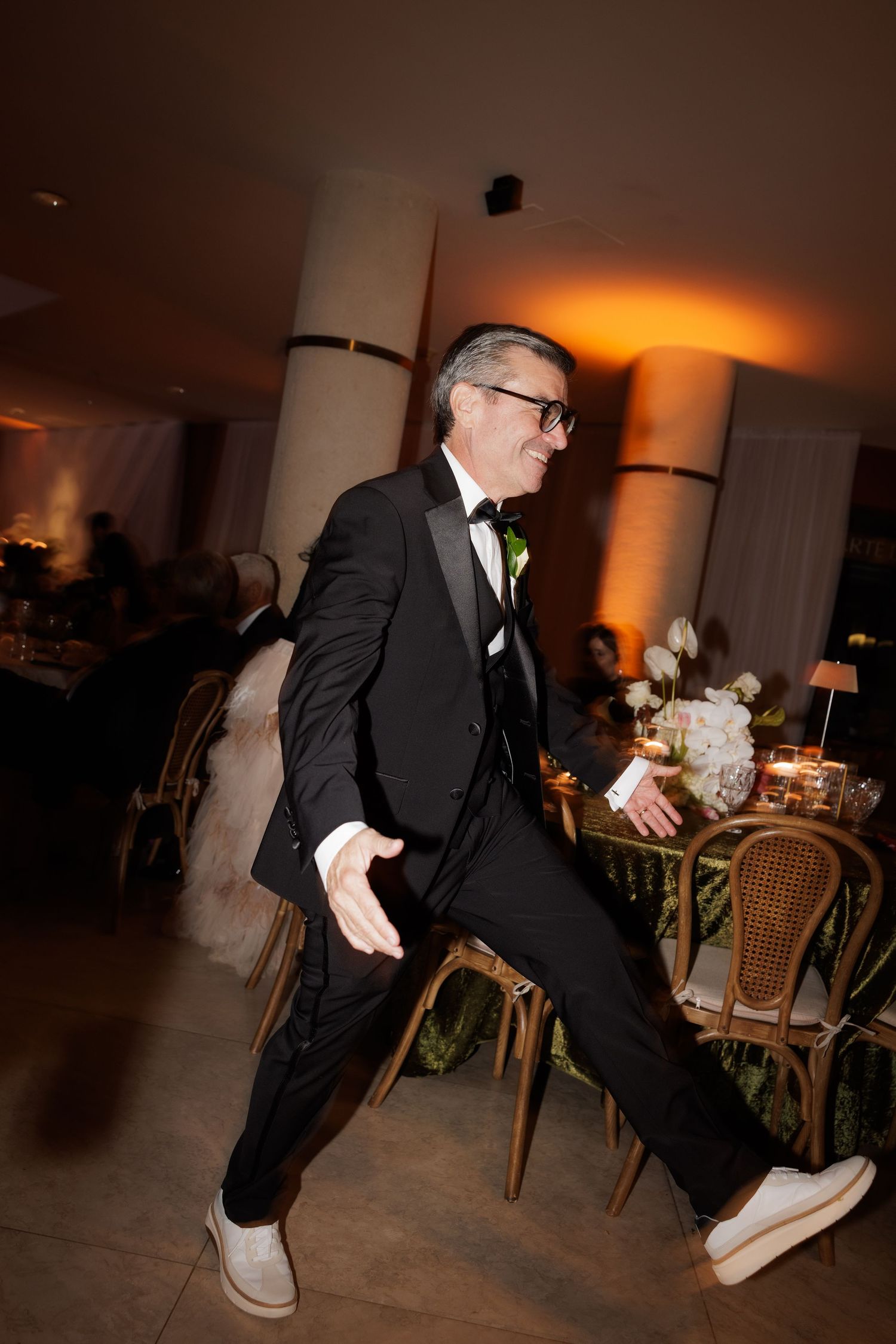 A man in formal attire performs energetic dance moves on a dimly lit reception dance floor.