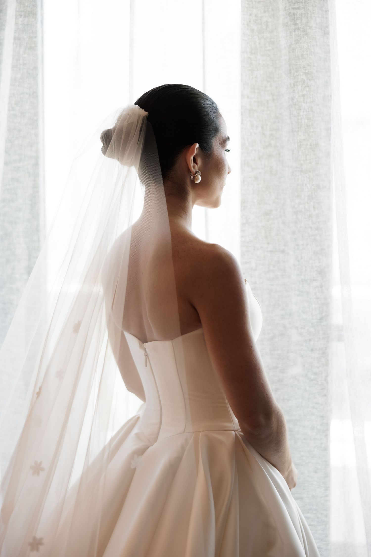 A bride in a white wedding dress and veil stands silhouetted against sheer curtains in soft natural light.