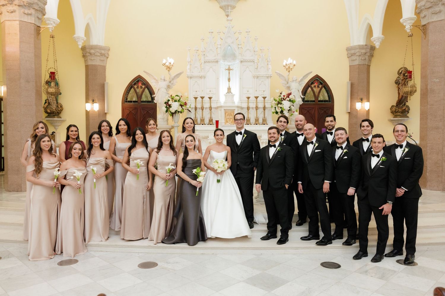 A formal wedding party poses in a church with bridesmaids in blush gowns and groomsmen in black tuxedos.