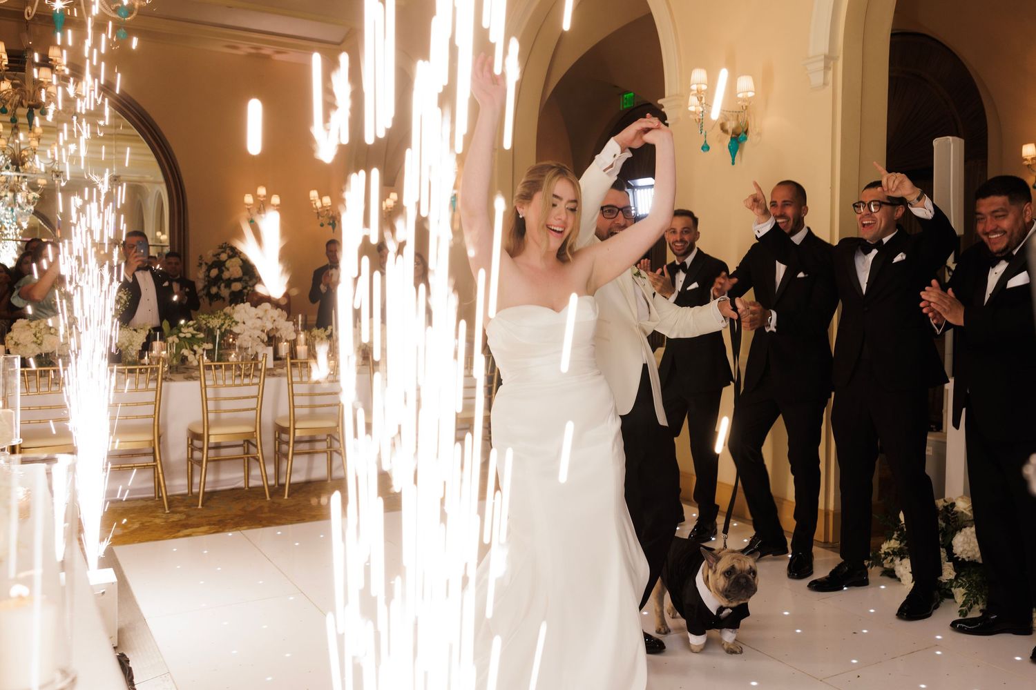 Wedding guests dance and celebrate on the dance floor during an elegant reception with chandeliers and warm lighting.