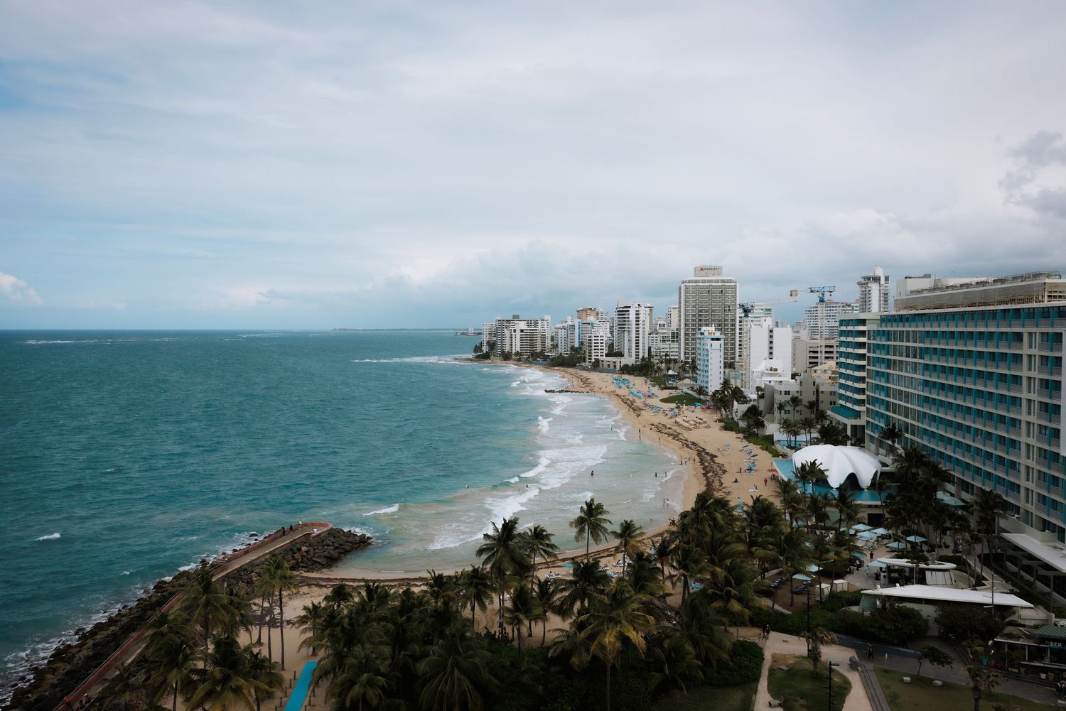 Aerial panoramic view of a coastal city skyline with modern buildings and beachfront stretching along turquoise waters.
