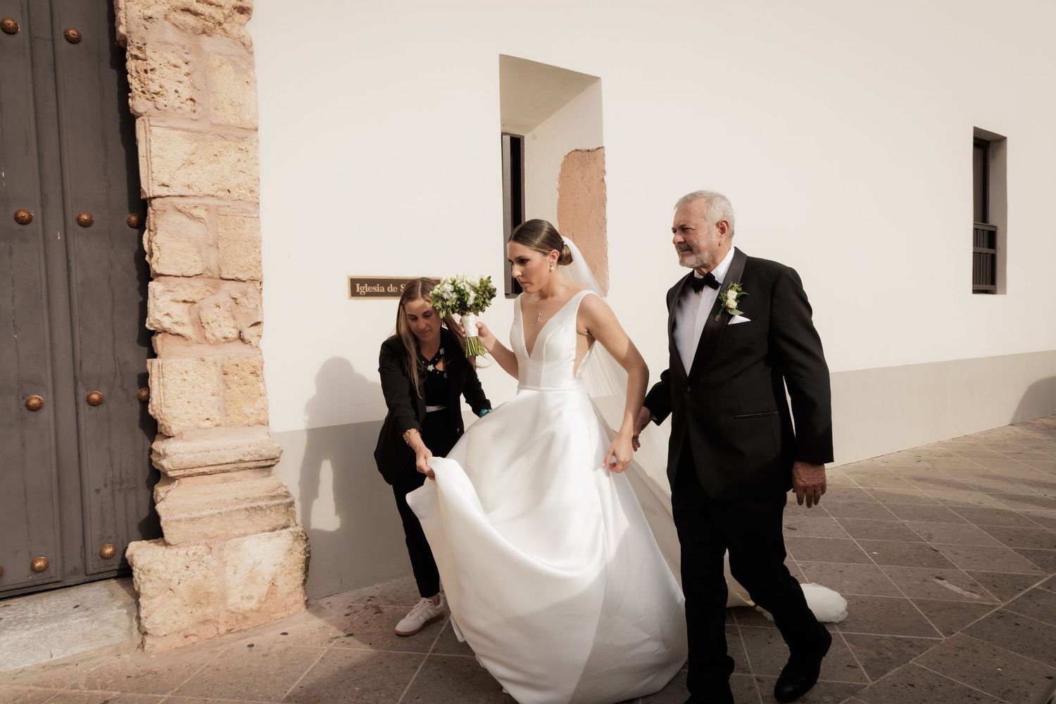 A bride in a flowing white wedding dress walks down a stone corridor with two people assisting her.