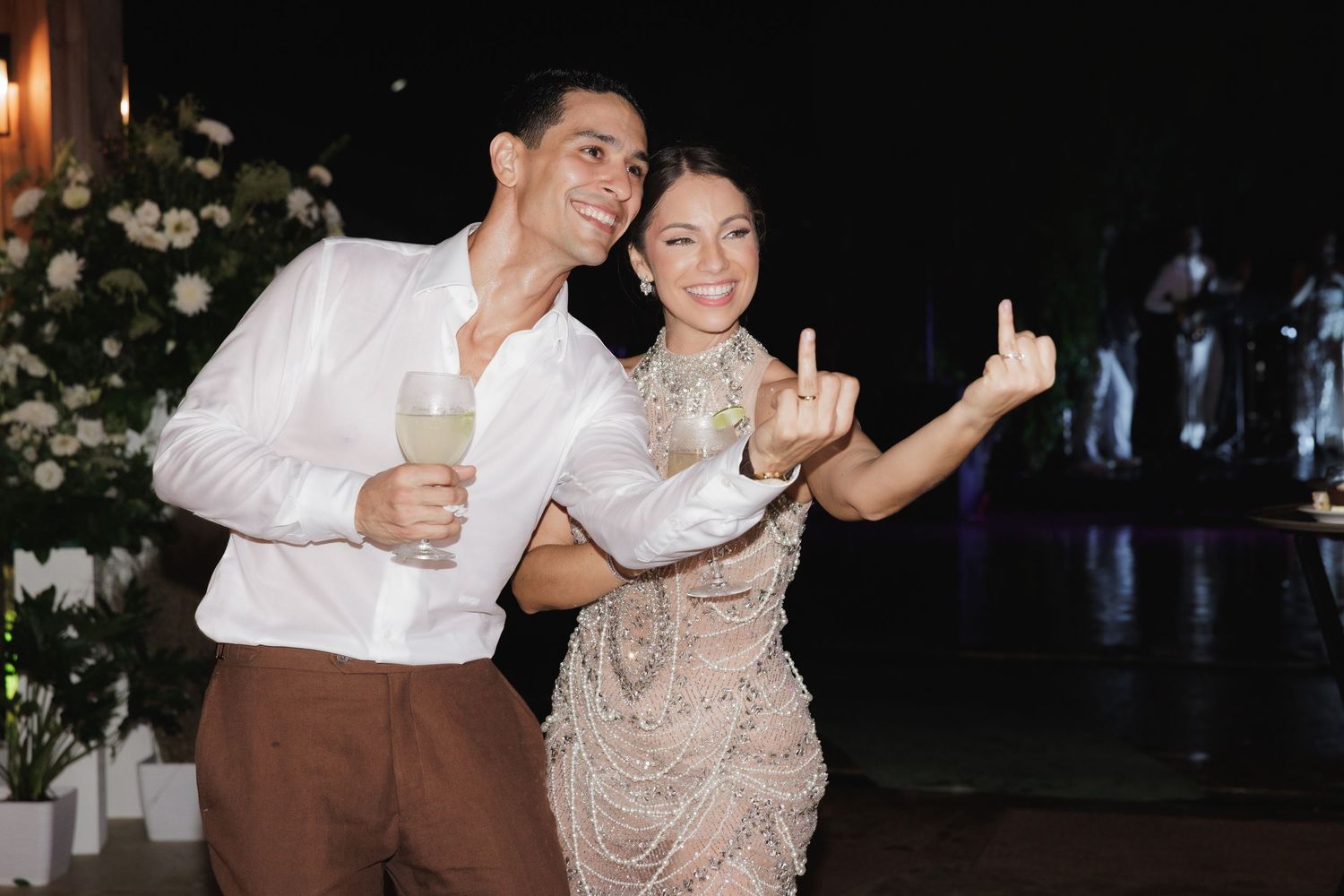 Two people dancing together at a nighttime celebration, with the person in a white shirt and brown pants holding a drink.