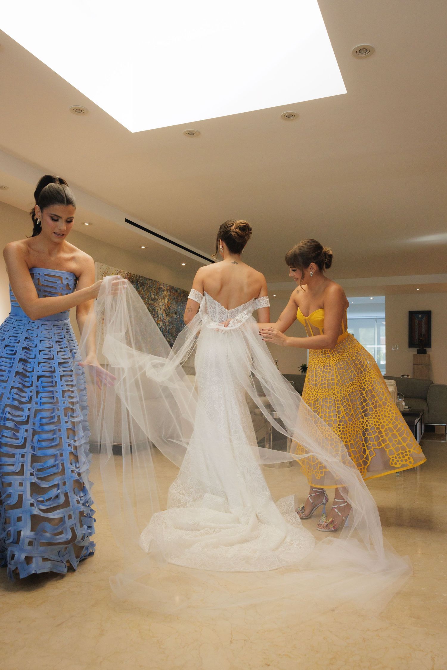 Bridesmaids in blue and yellow dresses help arrange the bride's wedding dress train indoors.