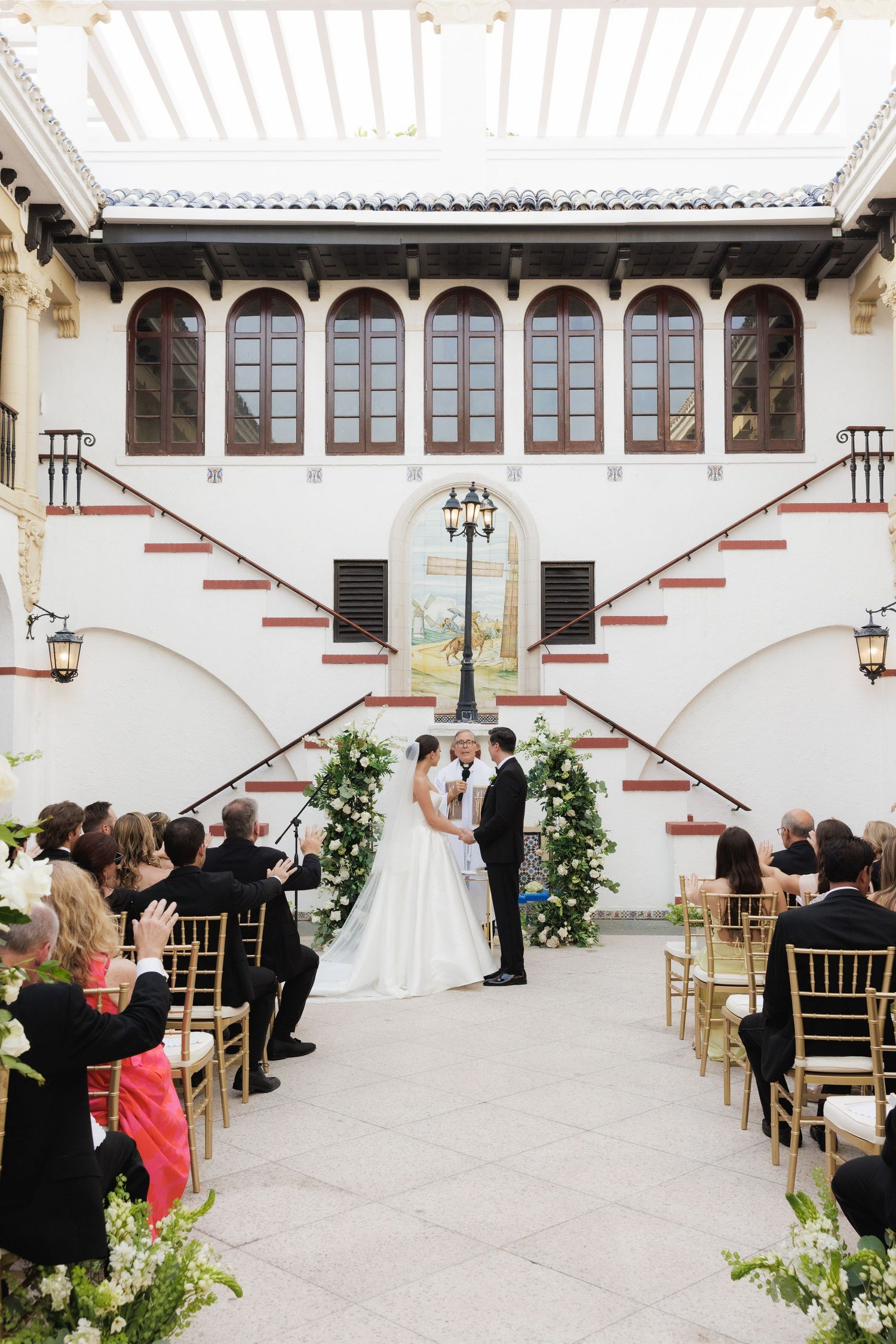 Outdoor wedding ceremony space with white columns and gold chairs arranged in rows.