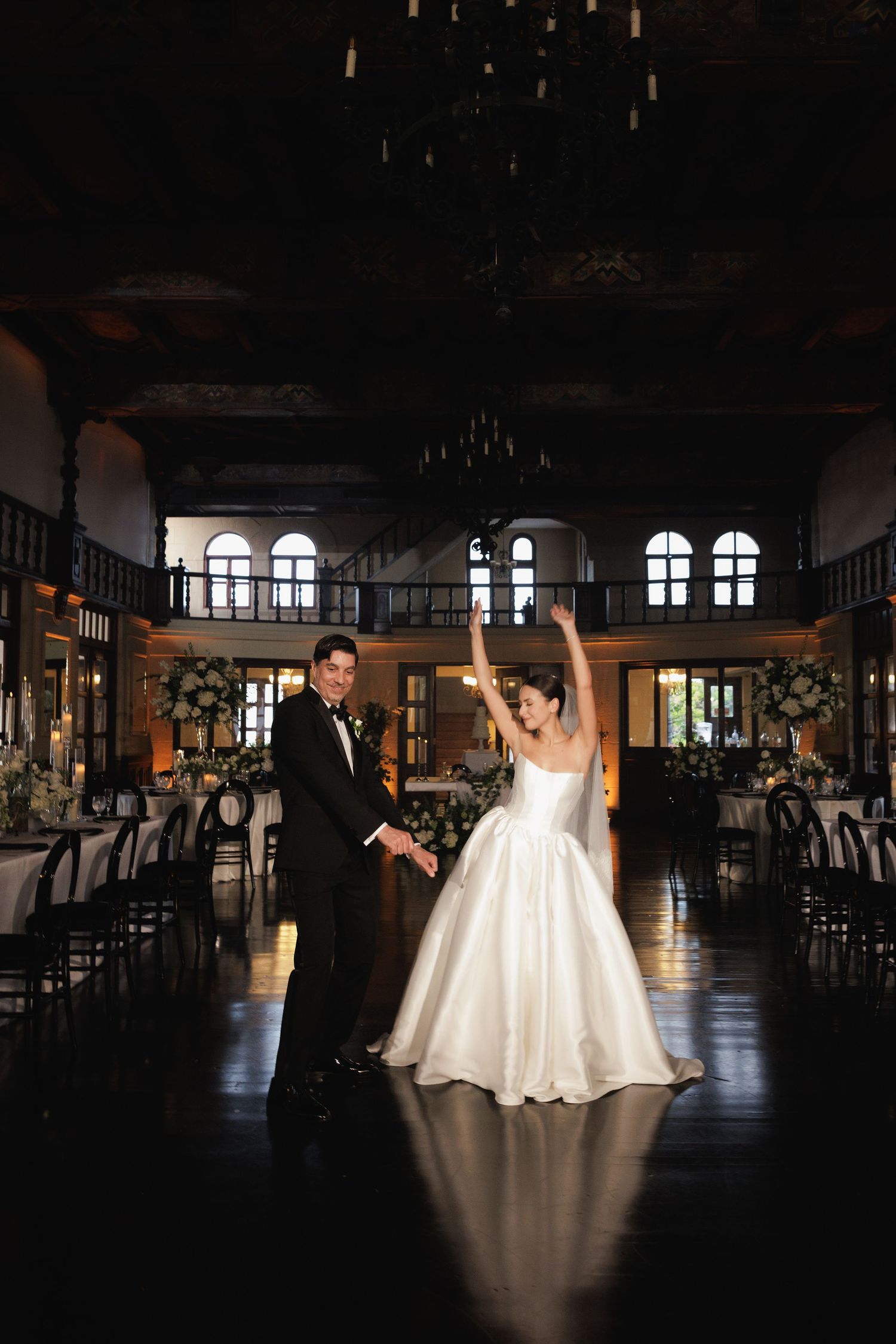 A couple dances in silhouette during their first dance in a grand ballroom with arched windows and dramatic lighting.