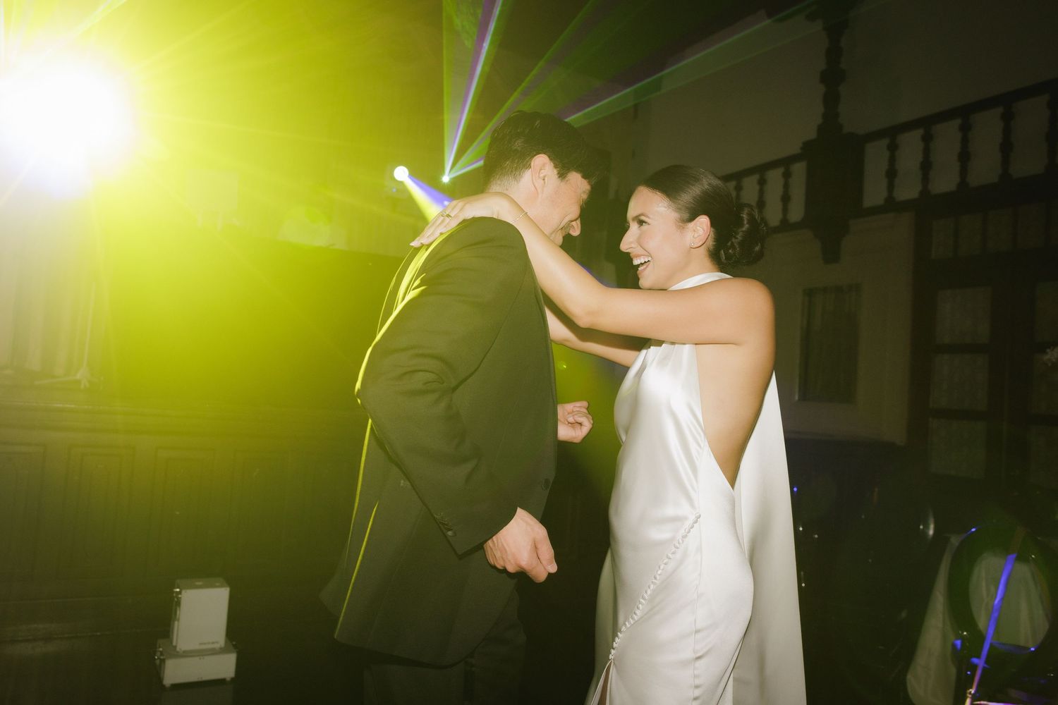 Wedding guests dance and celebrate on a dimly lit dance floor illuminated by yellow-green lighting.