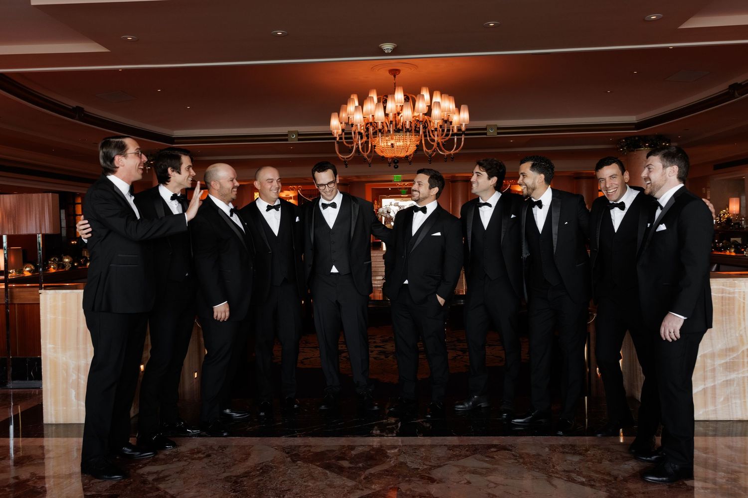 A group of formally dressed people stand together during a wedding ceremony in an elegant ballroom with chandeliers.
