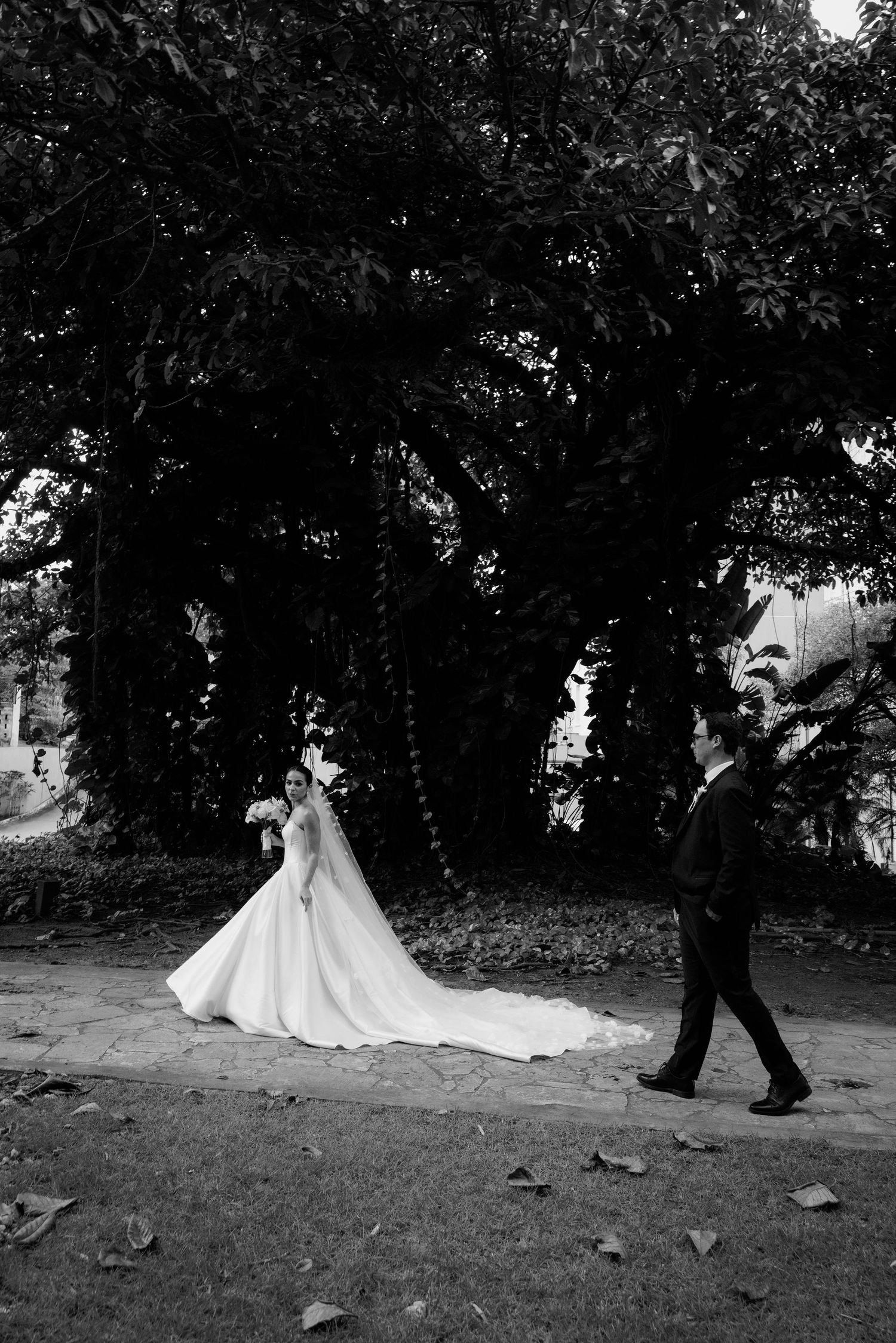 A black and white photo of a couple in wedding attire beneath dramatic tree coverage.