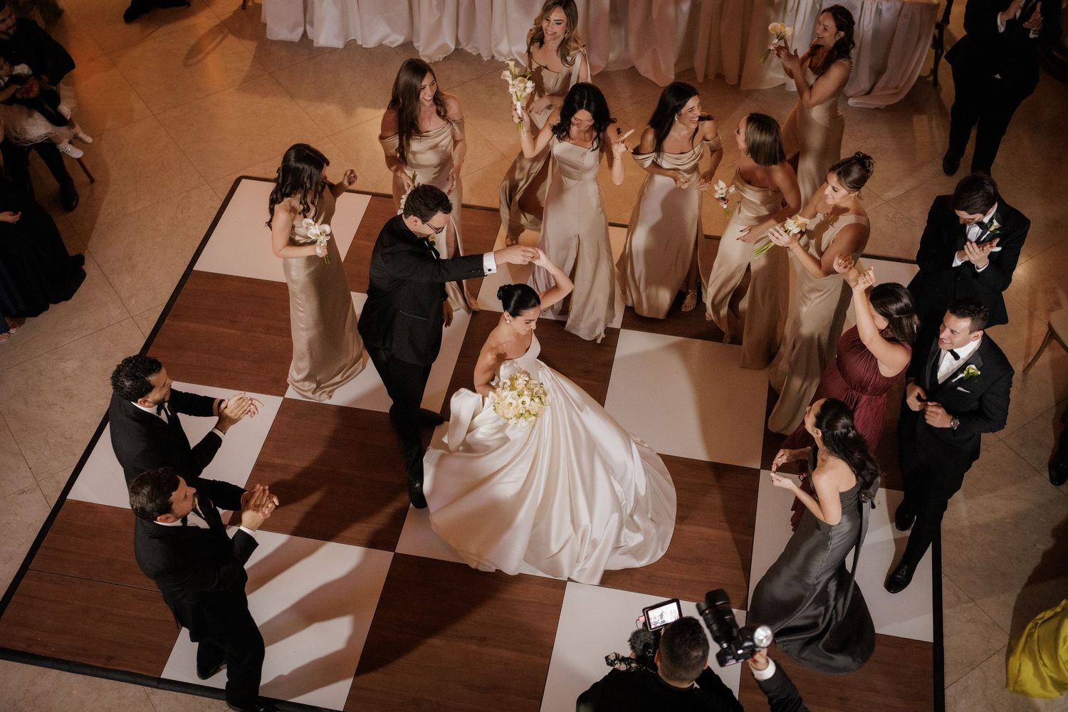 Wedding couple performs their first dance surrounded by guests with cameras on a wooden dance floor at reception.