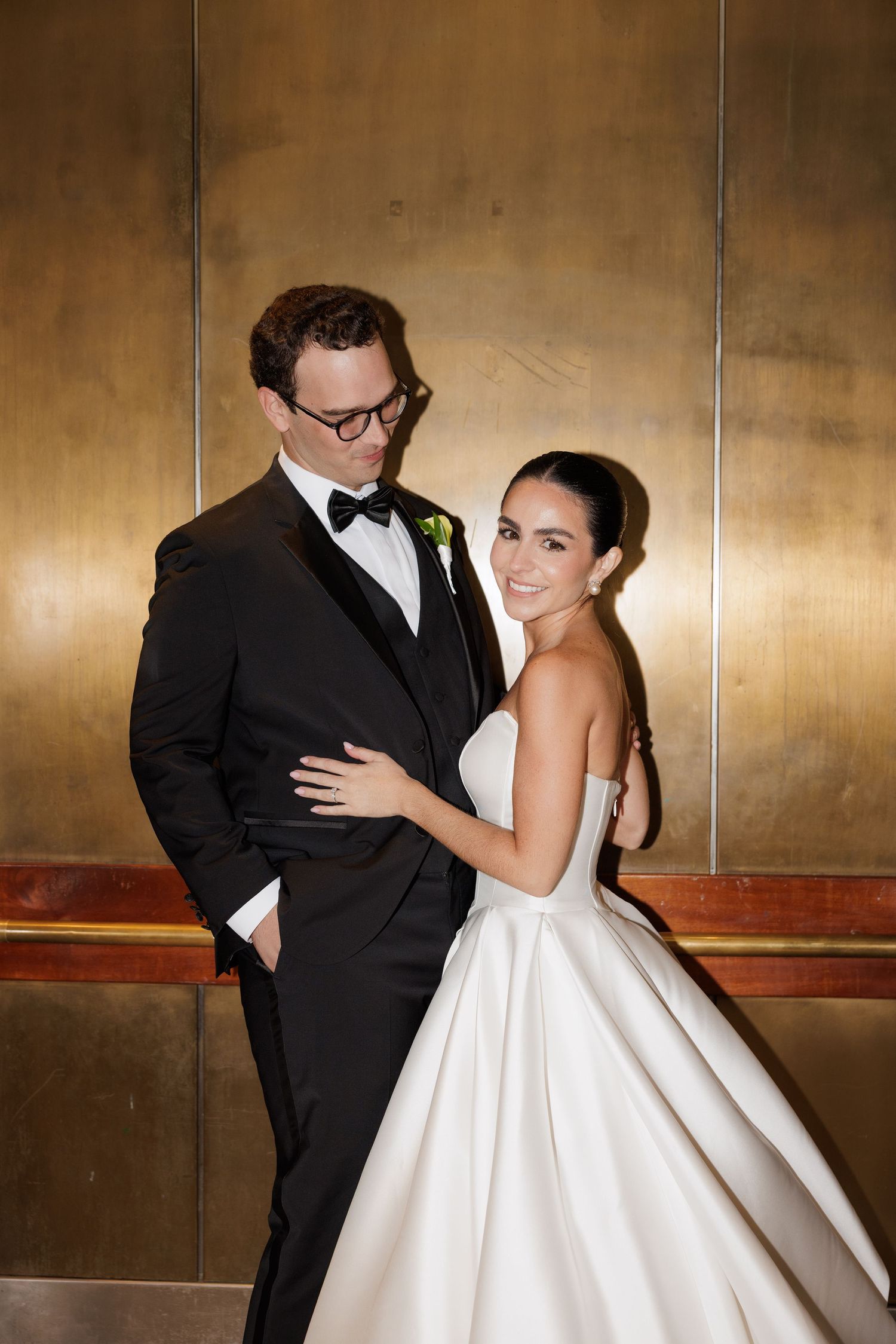 Wedding couple poses together against warm golden metallic wall in elegant formal attire.