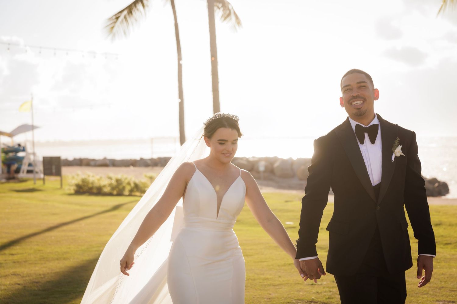 A newlywed couple walks hand in hand on a beautiful golf course with palm trees and sunlight in the background.
