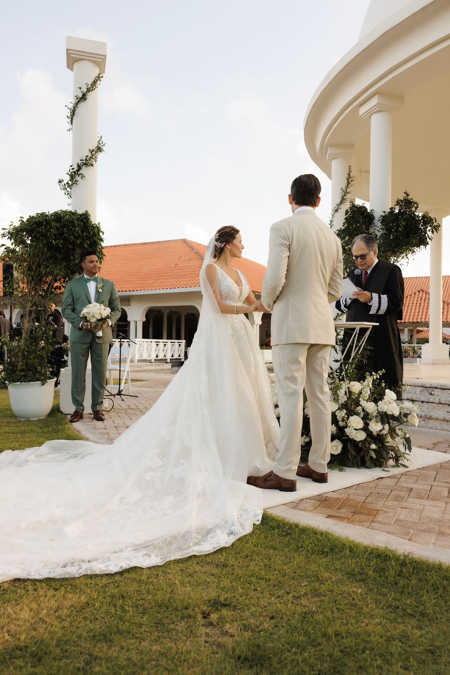 A bride in a flowing white gown with a long train poses on a porch with arched columns at sunset.