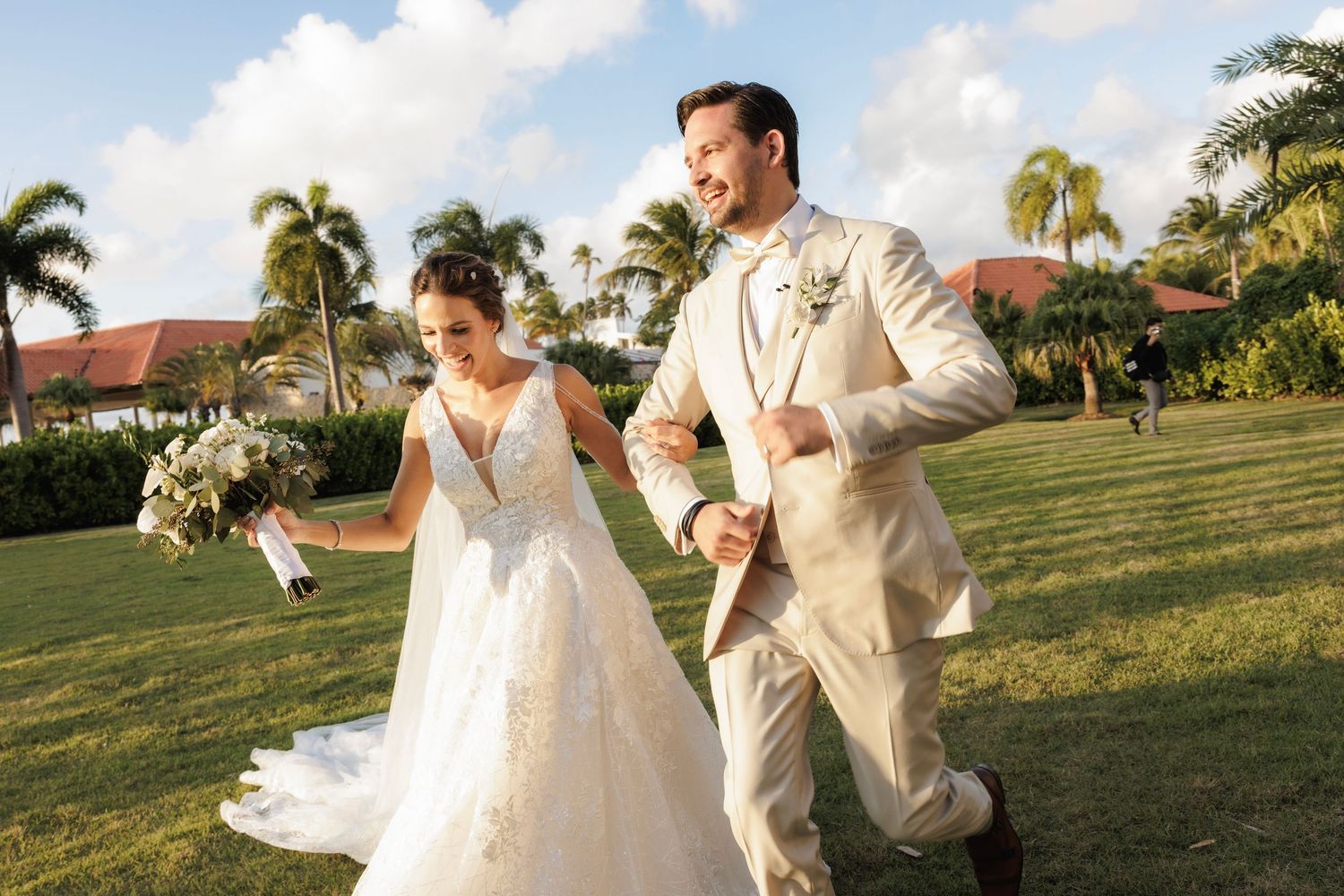 A couple in wedding attire dance playfully in a field at sunset with palm trees in the background.