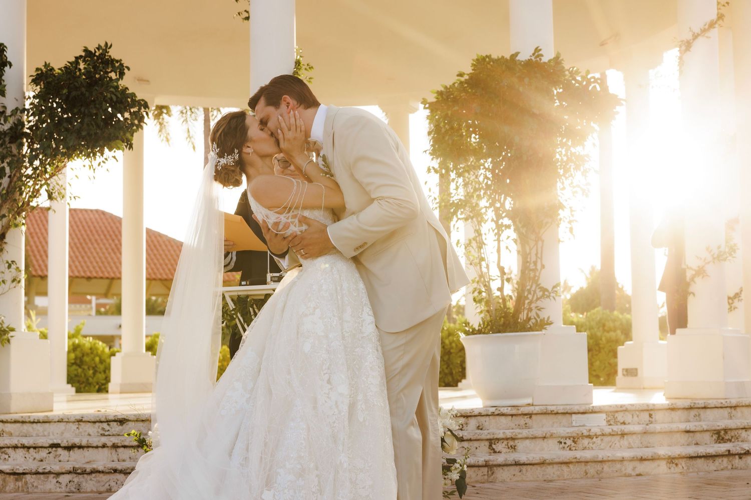 Bride and groom share a romantic kiss during golden hour on grand white pillared steps at a luxury wedding venue.