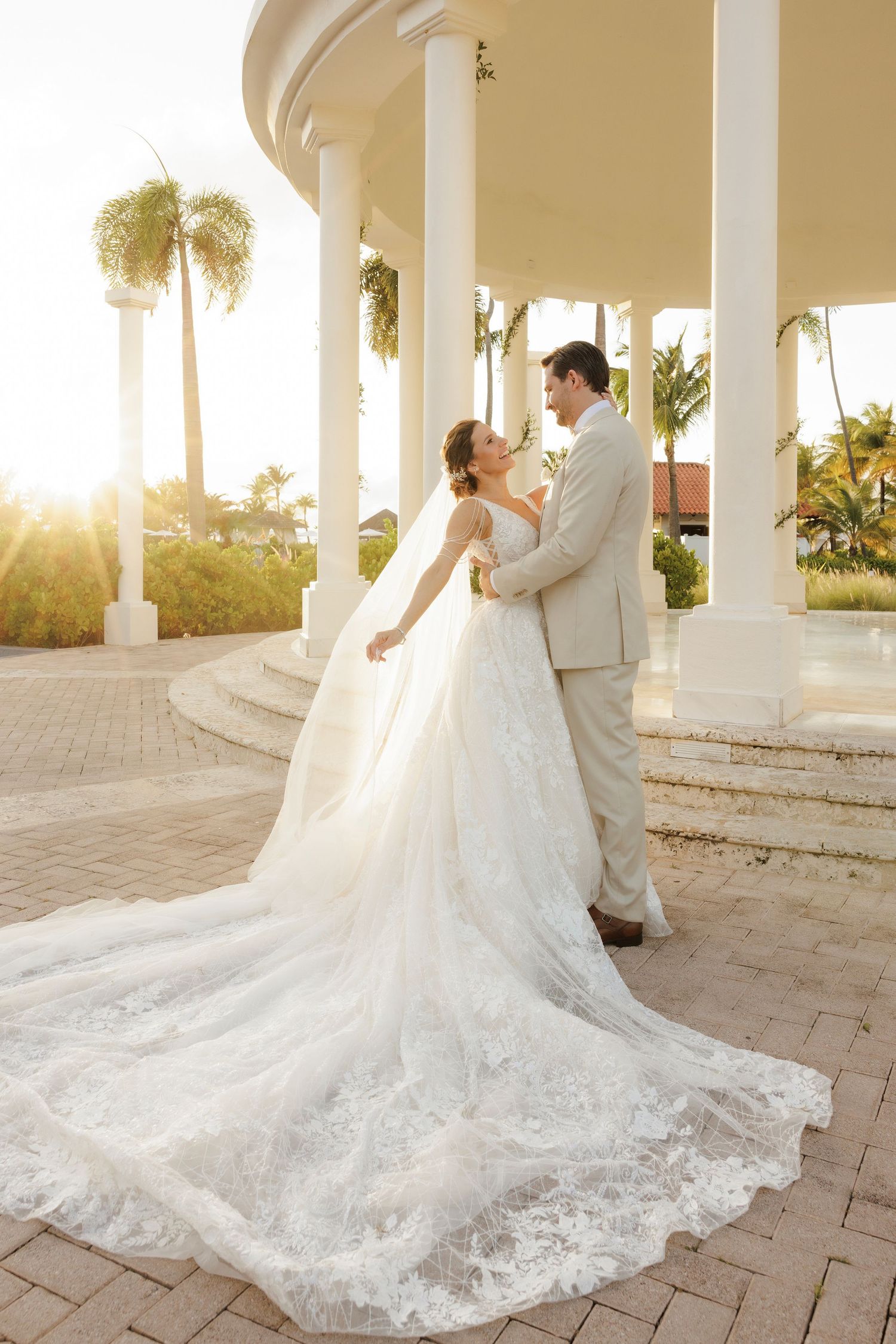 A bride in an elegant lace gown with dramatic train poses under white columns at sunset during a wedding photoshoot.