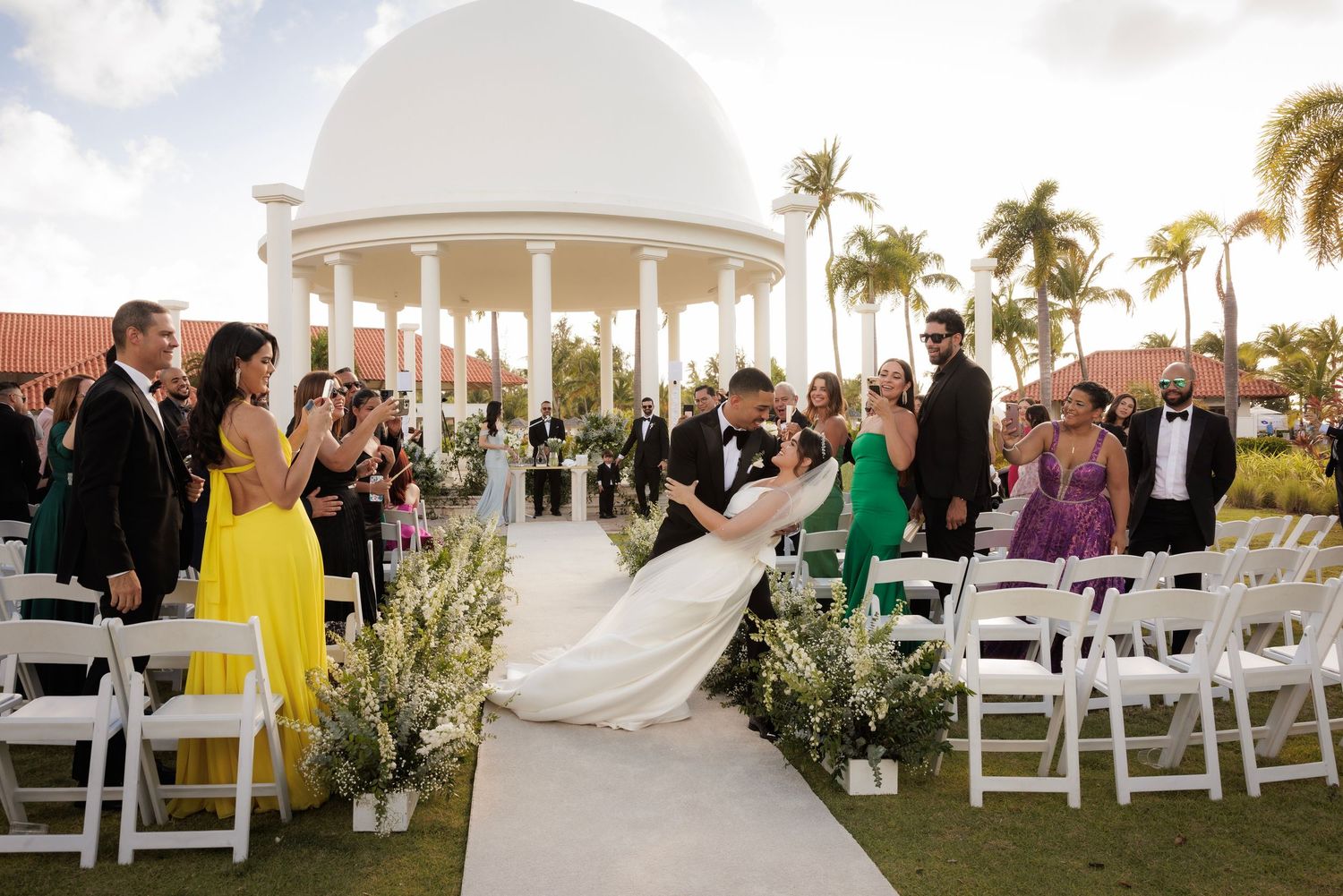 A wedding ceremony setup on the beach with white chairs, arch and palm trees at sunset.
