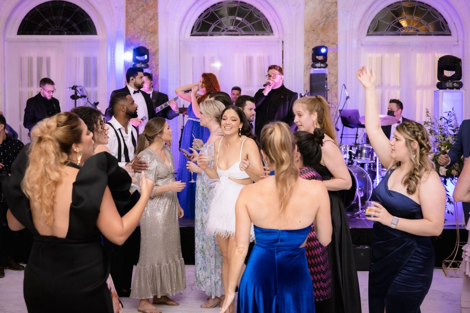 Couples dance together during a wedding reception in a ballroom with purple uplighting and arched windows.