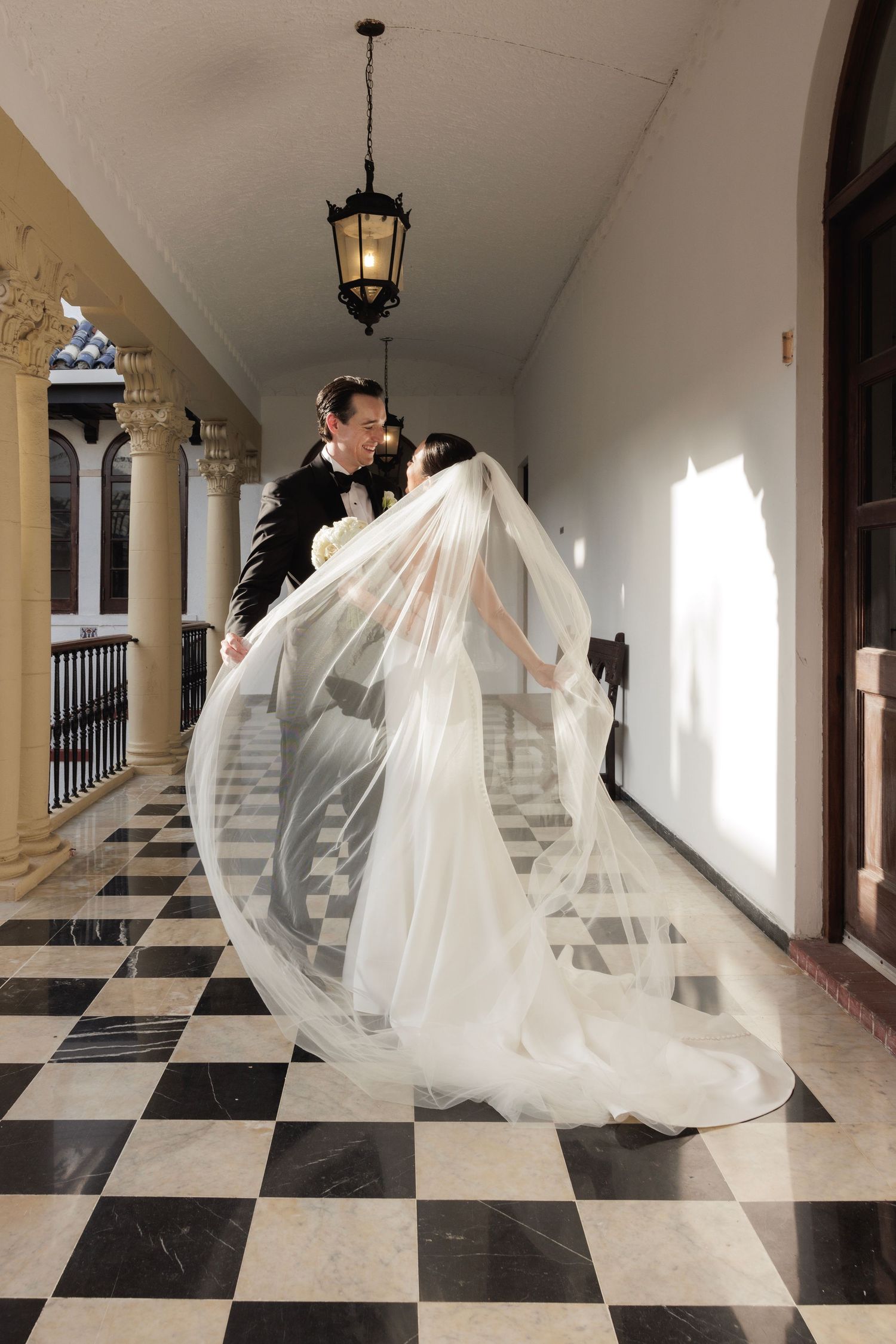 Wedding couple creates flowing movement with bride's veil in a classical colonnade with checkerboard flooring.