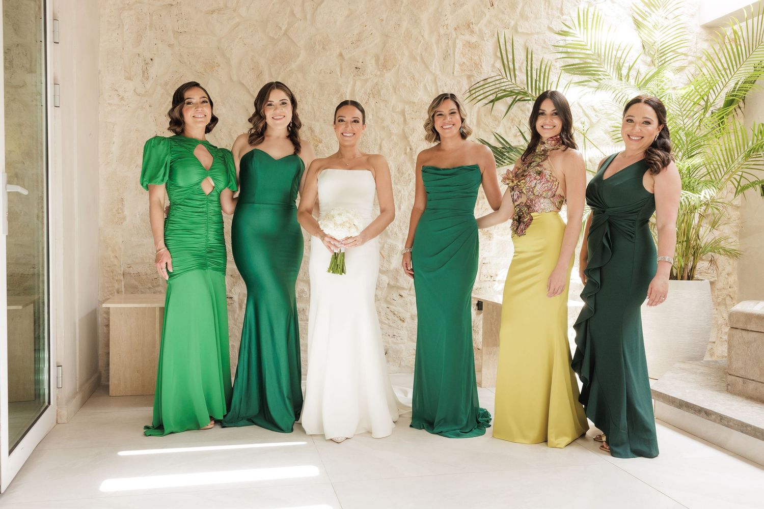 Wedding party members in coordinated emerald green dresses pose together with bride in elegant white gown.
