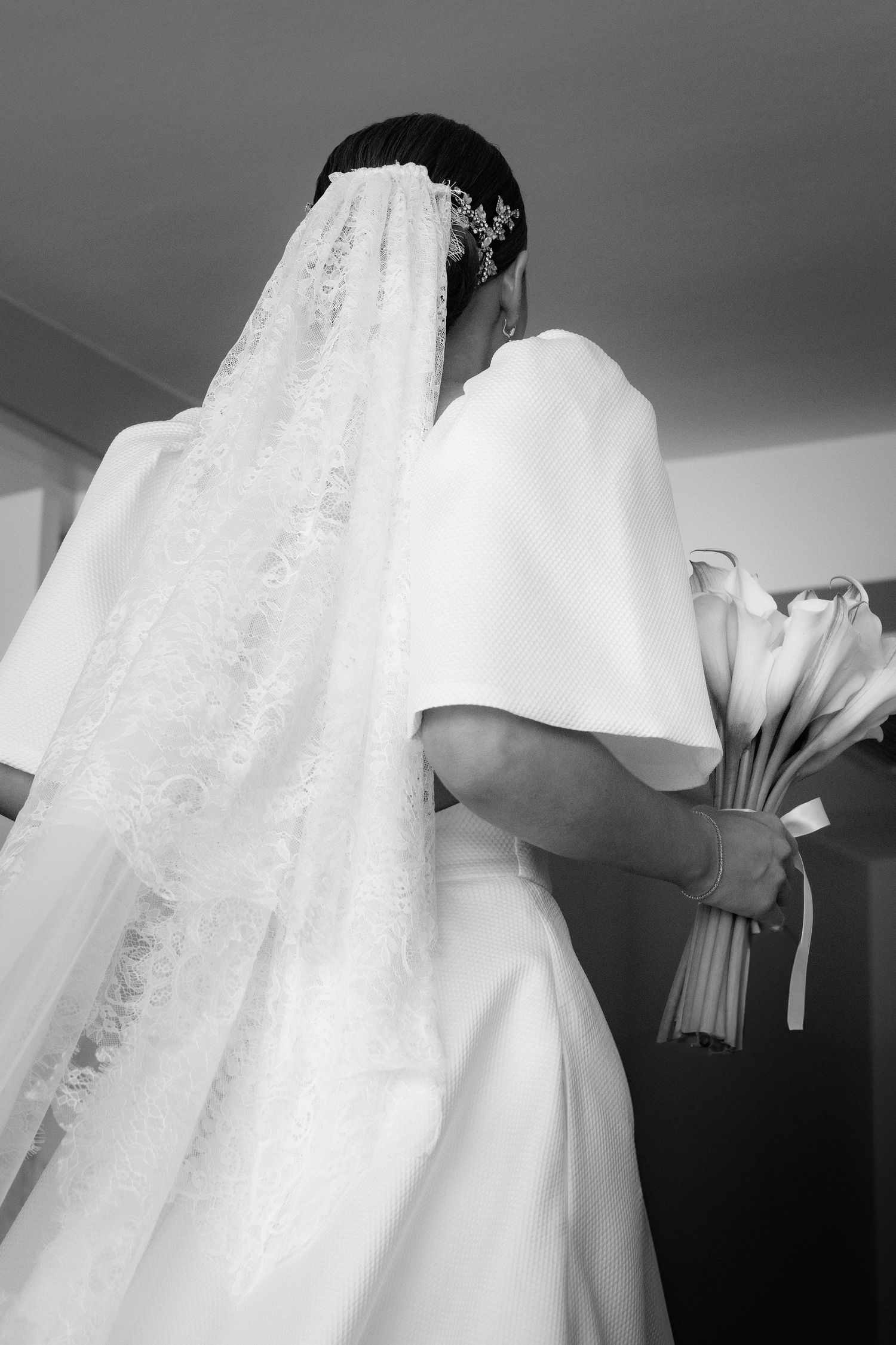 A bride in an elegant white dress with a long lace veil holds a bouquet in a black and white photograph.
