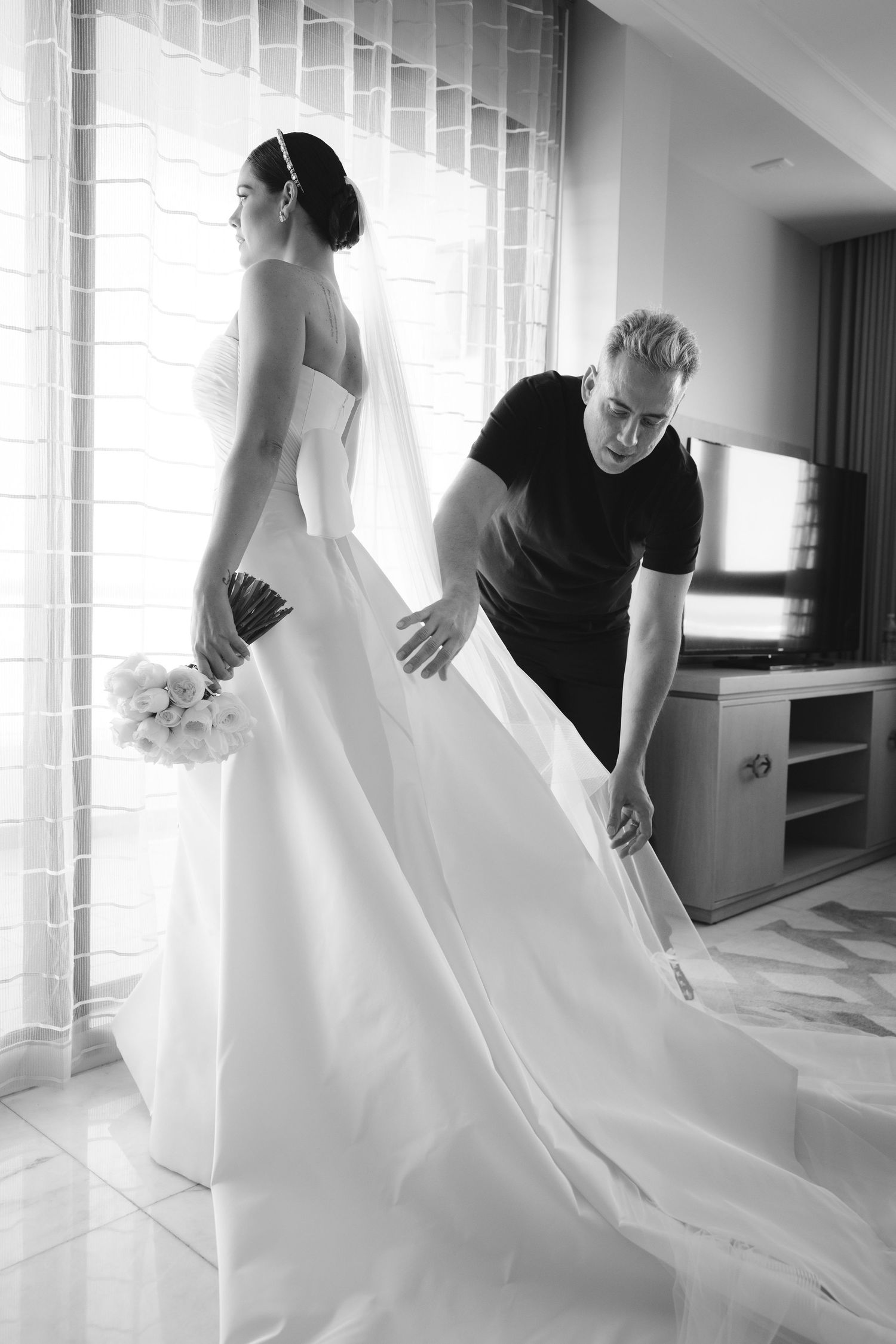 A person in a flowing white wedding gown stands by a window while someone adjusts the dress train and veil.