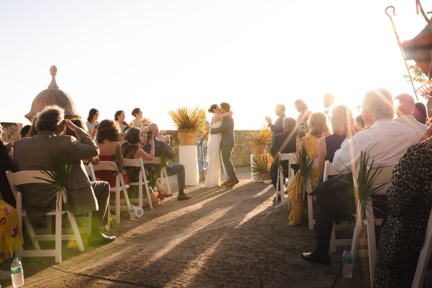 Wedding guests gather outdoors at sunset creating long shadows across a scenic venue.