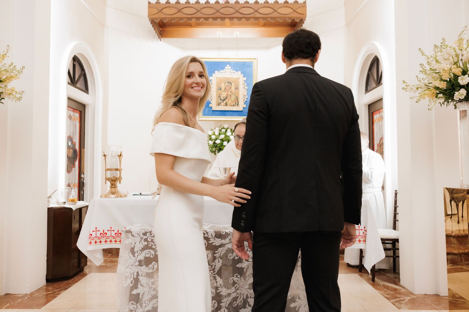 A bride and groom walk through an elegant white hallway decorated with ornate gold accents and floral arrangements.
