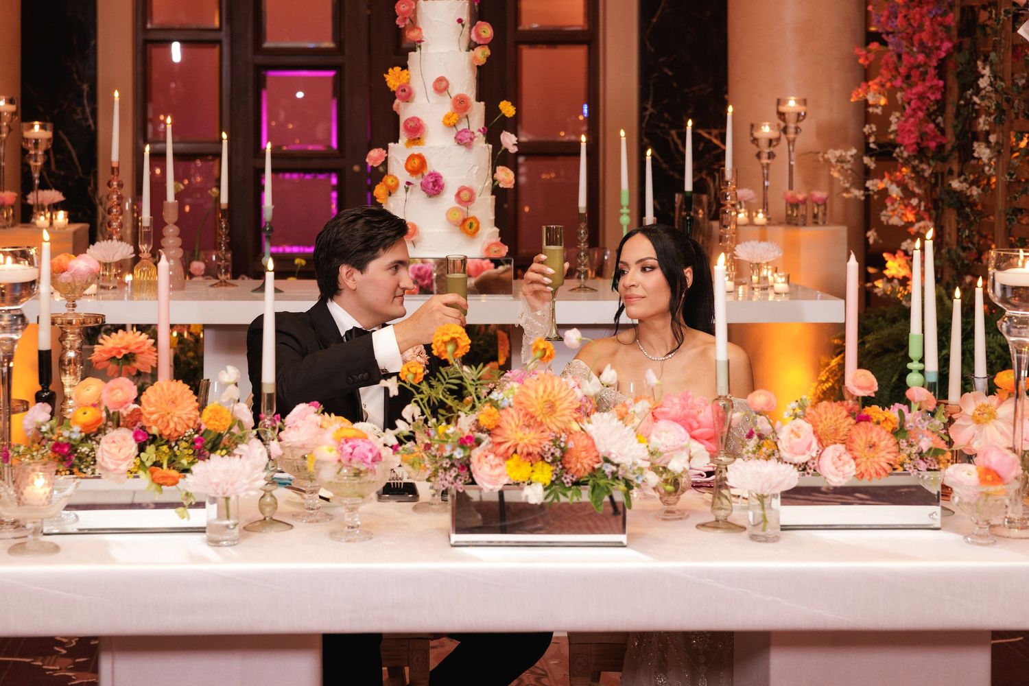 Elegant wedding reception table decorated with peach and coral flowers, tall candles and a tiered white cake in the background.