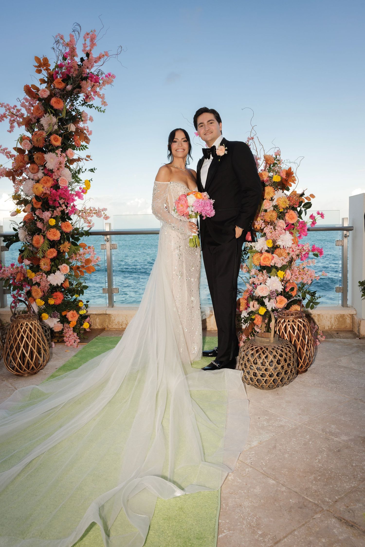 A wedding couple poses under a stunning floral arch with coral and pink blooms against an oceanfront backdrop.