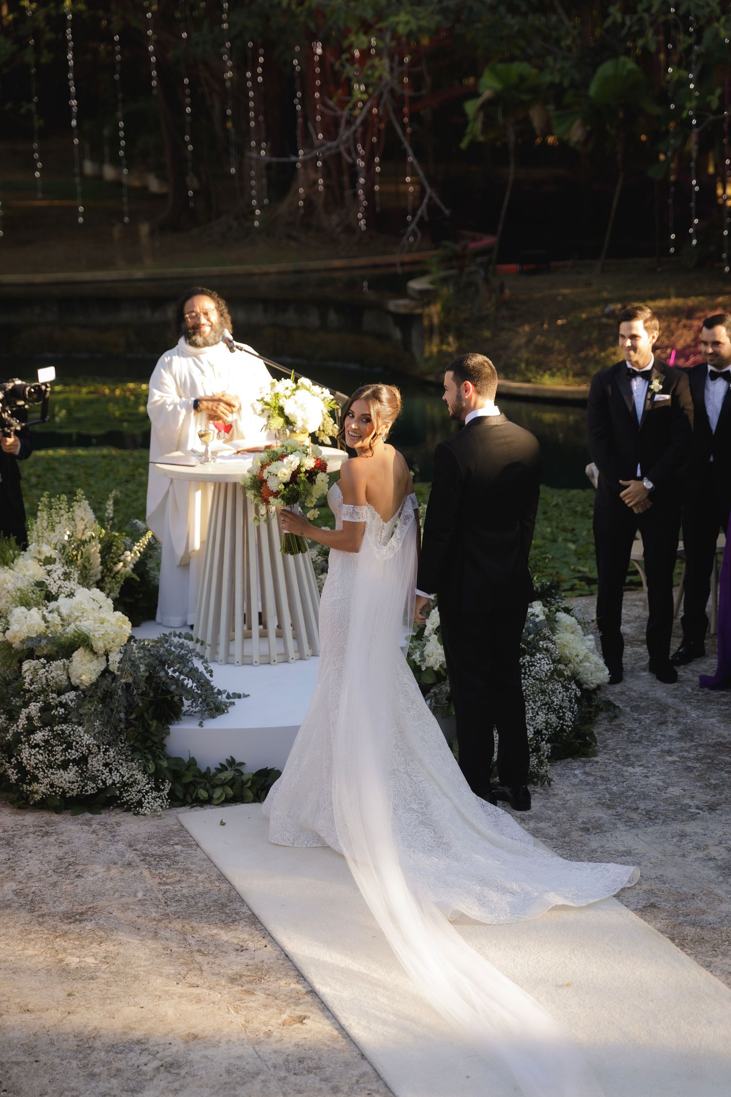 An evening wedding ceremony with string lights and white floral decorations along a garden path.