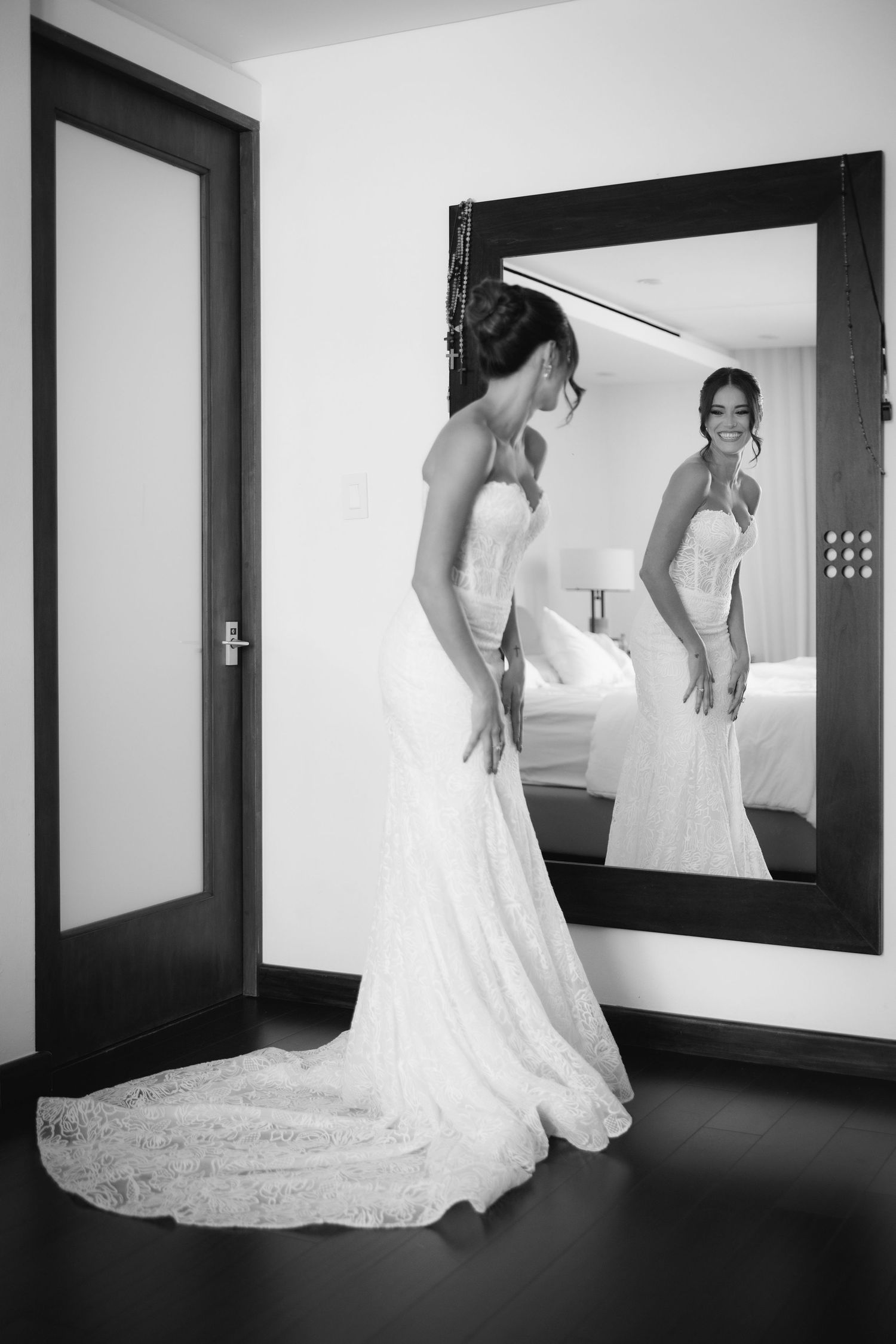 Bride in elegant lace wedding gown admires her reflection in a full-length mirror in black and white photo.