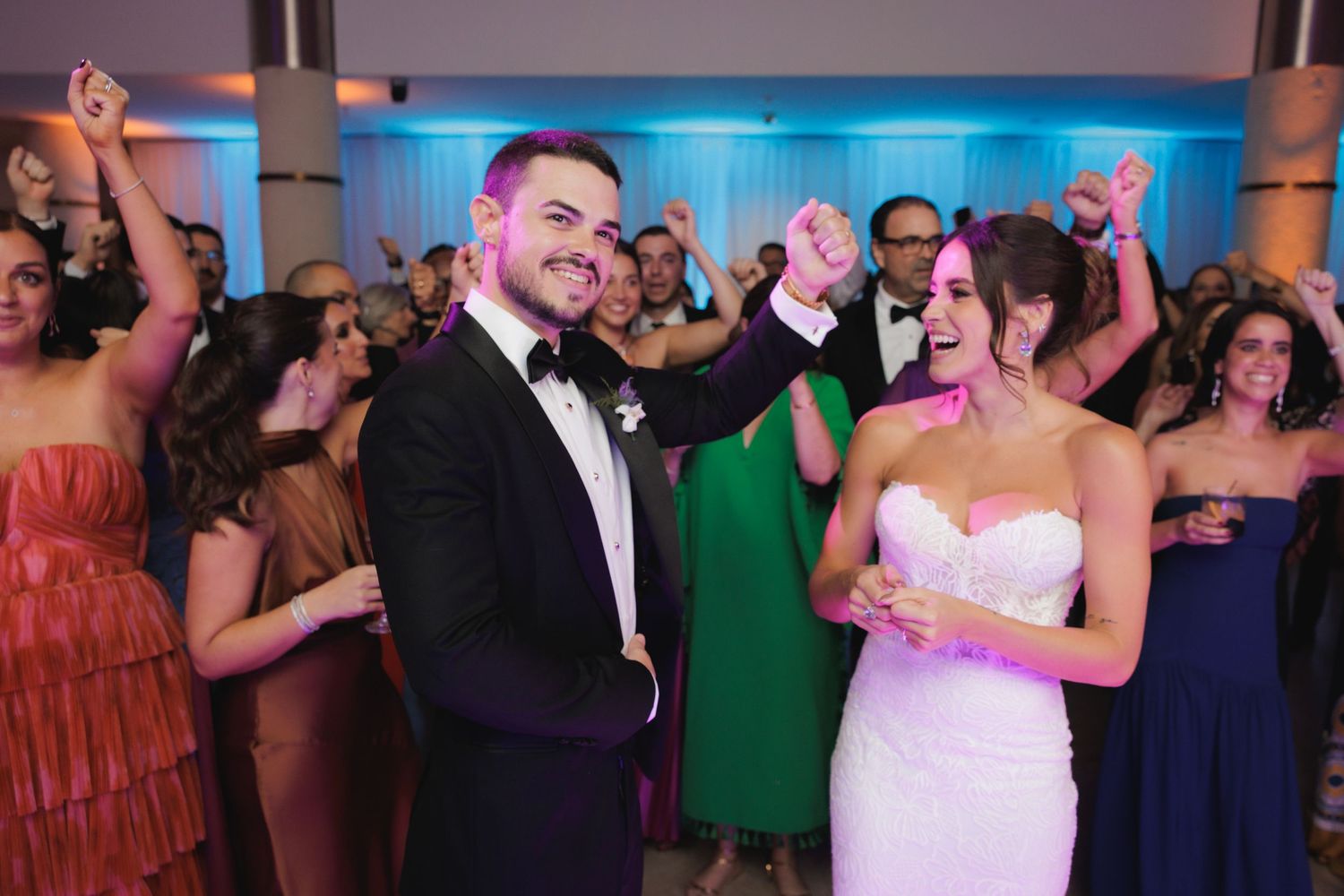 Wedding guests celebrate and dance together at an elegant reception with blue lighting and formal attire.