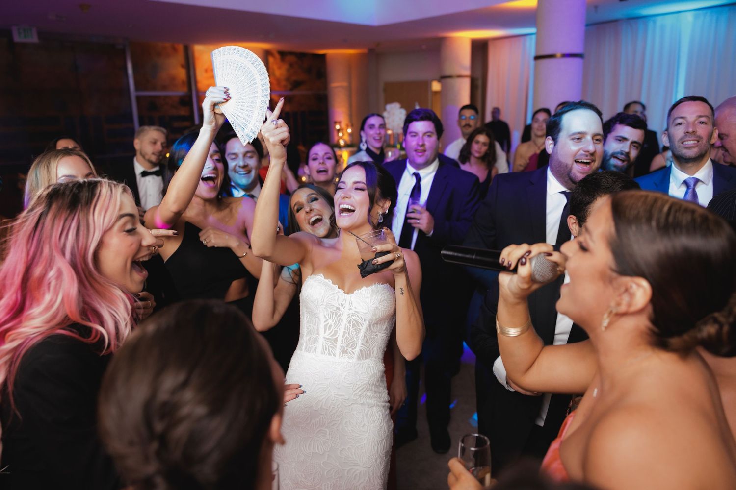 Party guests singing and celebrating on a crowded dance floor at a wedding reception with purple mood lighting.